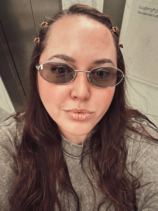 A close-up selfie of a woman with long dark wavy hair wearing tinted oval sunglasses and copper-colored hair clips, wearing a gray ribbed top in what appears to be a bathroom setting.