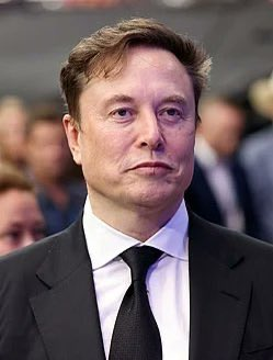 A photo of Elon Musk in a dark suit and black tie, appearing at what looks like a public event with a crowd blurred in the background. The image relates to the post by highlighting Musk, whose father claims he and his brother collectively hold 23,000 Bitcoin valued at over $1.776 billion.