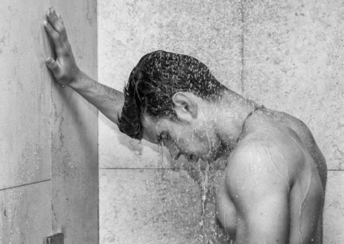 There are two people shown in the image - one person standing under a shower head with their arms resting against the wall while another person stands further away from them.