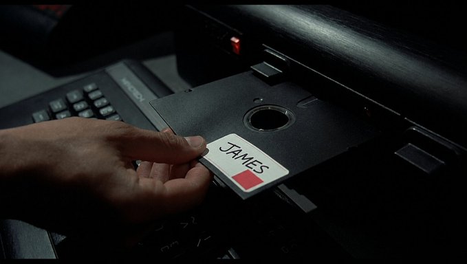 A hand places a 3.5" floppy disk labeled "JAMES" with a red square into a disk drive next to a MicroTouch keyboard.