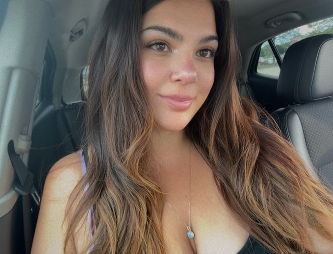 A woman with long, wavy brown hair sits in the passenger seat of a car, wearing a necklace with a turquoise pendant.