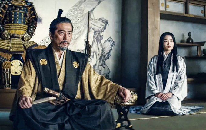 A man in traditional Japanese armor and robes sits with a sword, while a woman in a white kimono kneels respectfully behind him in a traditional room with samurai decor.