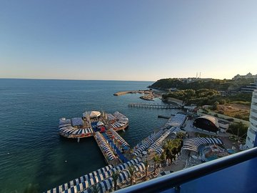 A scenic ocean view from a balcony, showing a calm sea, a small pier or jetty with covered lounging areas, and a tree-lined coast under a clear blue sky.