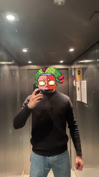 The image shows a man wearing glasses, standing inside an elevator with his face partially obscured by a mask that resembles a character from a video game or anime. The man appears to be taking a selfie using his cell phone while wearing the mask. The background of the image is a gray wall, and there are signs hanging on it, which could possibly be related to the gaming world or an event happening in the area.