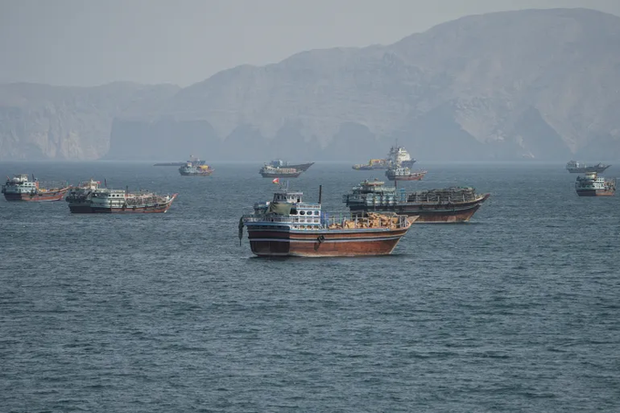 A fleet of wooden fishing boats and larger vessels floating on the water with hazy, rugged mountains in the background.