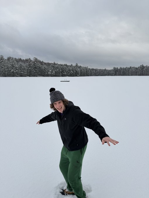 A young person wearing a gray beanie, black jacket, and green pants stands smiling on a frozen, snow-covered lake with a forested shoreline in the background.