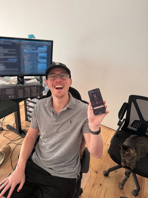 A young man wearing glasses and a cap sits at a desk with computer monitors behind him, smiling and holding up his smartphone to display an app screen, with a tabby cat sitting on an office chair beside him.