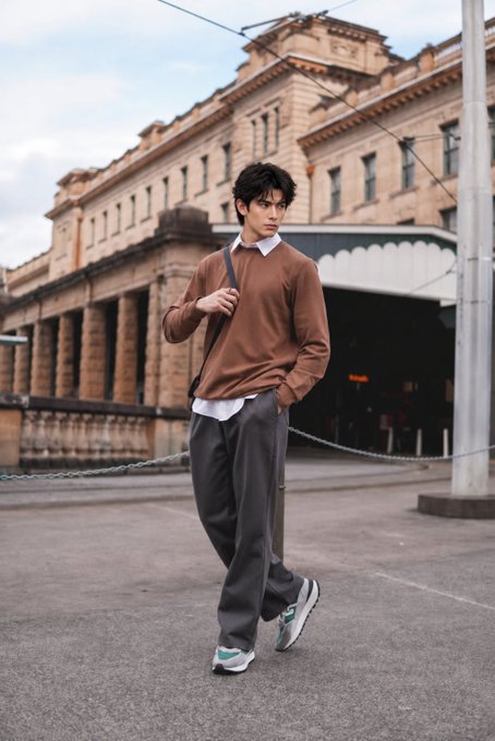 A young man with dark hair walking down a street, wearing a brown sweater over a white collared shirt, gray pants, and sneakers, with a large historic stone building in the background.