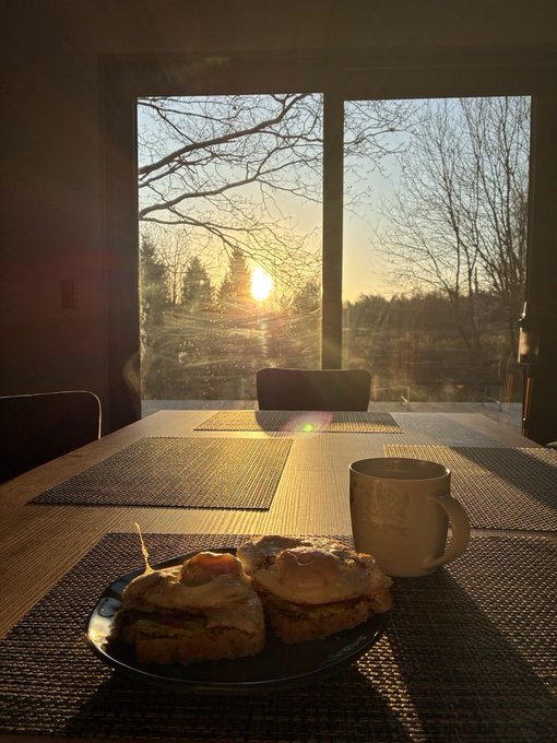 The image shows a breakfast scene with two sandwiches placed on a plate next to a coffee cup. The setting appears to be indoors, possibly in a dining room or kitchen area, as there are chairs around the table. A window can also be seen in the background, providing natural light and a view of trees outside. The post suggests that small daily actions, like consistently showing up for breakfast and learning from it, can lead to significant personal growth over time.