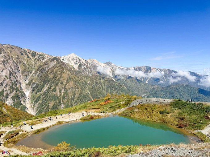 A scenic mountain landscape featuring a turquoise alpine pond in the foreground and rugged mountain peaks with low-hanging clouds in the background, with small figures of hikers along the trail.