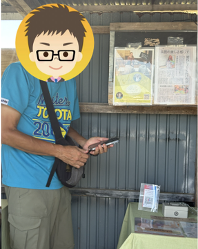 A person in a blue Toyota t-shirt uses a smartphone to scan a JPYC QR code at an unmanned green onion stall in Akita, demonstrating successful cryptocurrency-based payments at a rural vending setup.