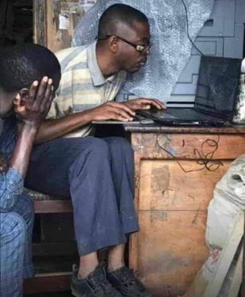 A young man wearing glasses sits at a wooden desk using a laptop, while another person partially visible in the foreground covers their face with their hand.
