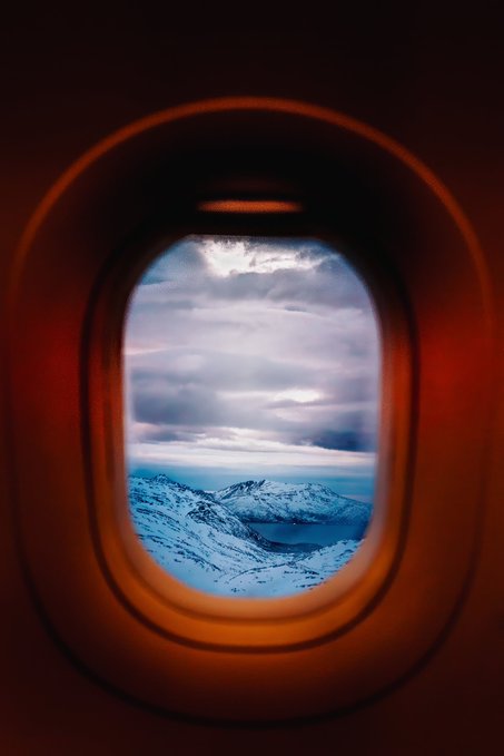 A view from an airplane window showing snow-covered mountains under a cloudy sky, framed by the warm orange glow of the cabin interior.