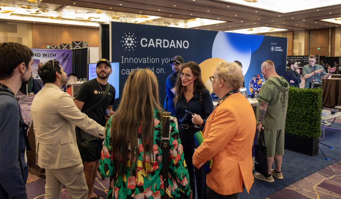 Attendees gather around the Cardano booth at the Rare Evo event, engaging in conversations in front of a branded backdrop reading "CARDANO Innovation you can build on." The booth serves as a popular networking hub, complementing the post's description of demos, governance discussions, and Ledger raffles.