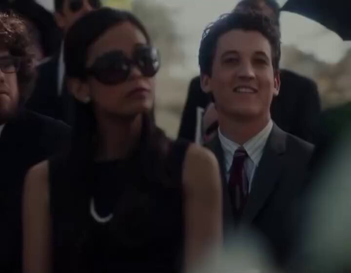The image shows a man wearing a suit and tie standing next to a woman dressed in black with sunglasses. They are both smiling at each other, creating an atmosphere of camaraderie or friendship. The background includes several people who appear to be engaged in conversation or observing something happening nearby. There is also an umbrella visible in the scene, suggesting that it might have been raining earlier and they are now gathered together for a social event.