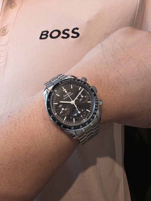 A close-up photo of a man's hairy wrist wearing a stainless steel Omega Speedmaster chronograph watch with a black dial, paired with a peach-colored Hugo Boss shirt.