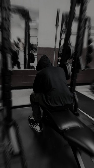 A moody black and white photo of a person in a dark hoodie sitting on a gym bench surrounded by weightlifting equipment.