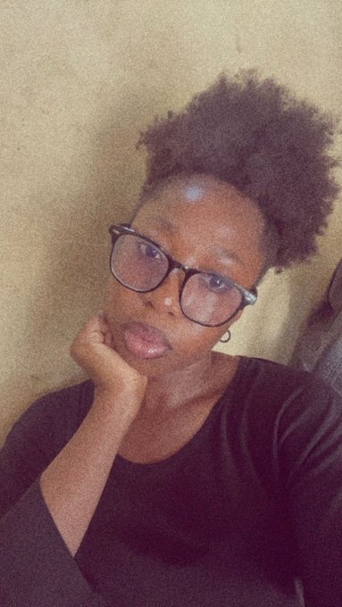 A woman with short natural hair and black-framed glasses rests her chin on her hand, looking at the camera in a selfie-style portrait.