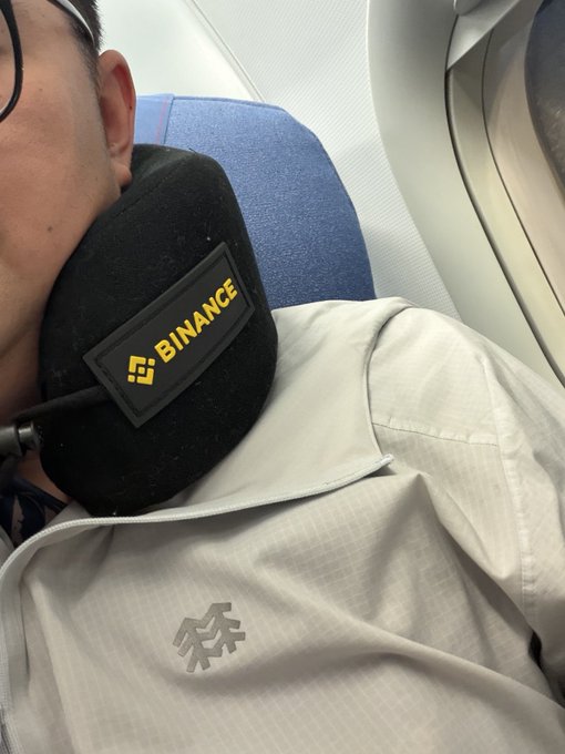 A close-up selfie of a person resting their head on a black neck pillow branded with the 'BINANCE' logo while on an airplane.