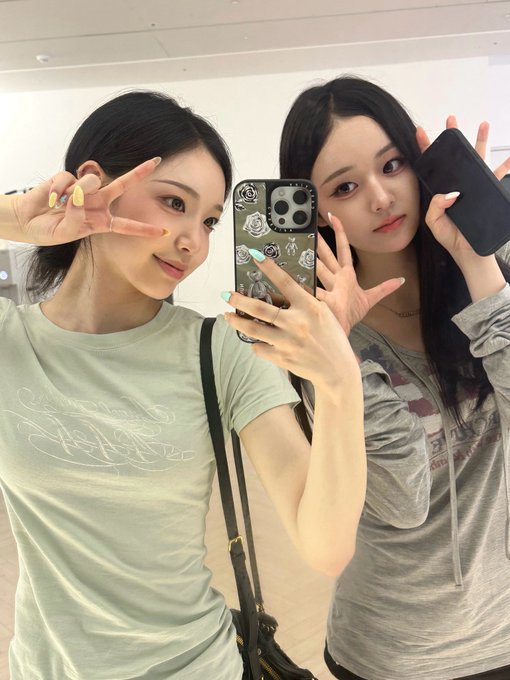 Two young women take a mirror selfie holding smartphones, both making peace signs, one wearing a green t-shirt and the other in a gray hoodie.