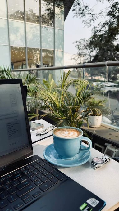 A blue cup of latte art coffee sits next to a laptop and wristwatch on a cafe table overlooking greenery and city streets.