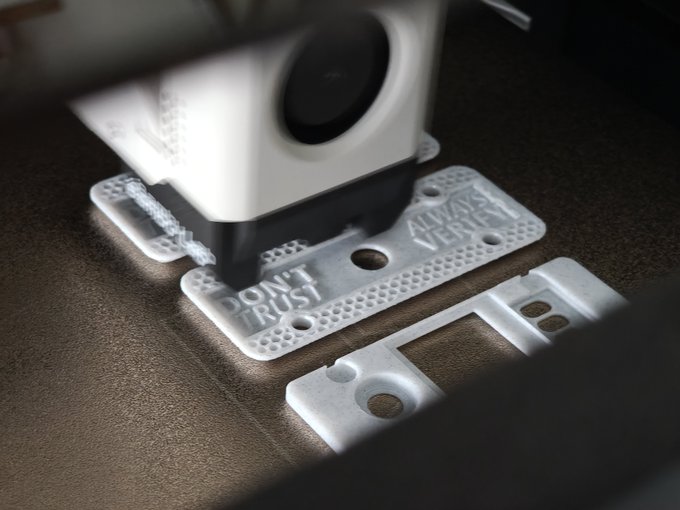A 3D printer extrudes white plastic to create small tag-shaped objects, with visible text reading "DON'T TRUST" and "ALWAYS VERIFY" on the partially completed prints.