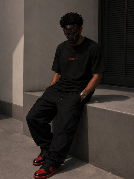 A person wearing a black samurai-style mask, a black t-shirt with red 'BASIT' text, black cargo pants, and red and black sneakers leans against a gray concrete ledge in a modern outdoor setting.