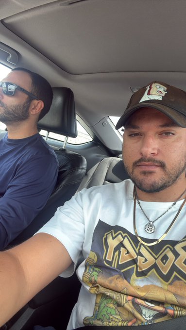 A selfie taken inside a car showing two men; one wears sunglasses and a blue shirt, while the other wears a baseball cap, chain necklace, and a 'KROK' graphic t-shirt.