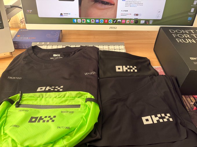 OKX-branded merchandise including a black t-shirt, hoodie, and neon green waist bag are laid out on a desk in front of a computer monitor.