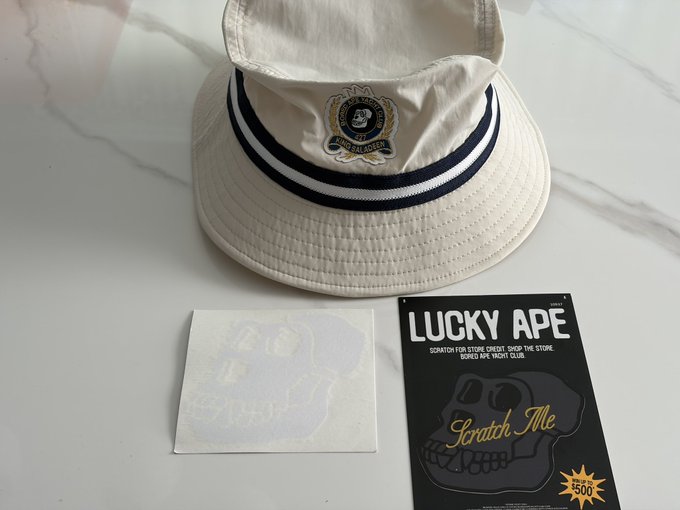 A cream-colored bucket hat with a 'LUCKY APE' emblem alongside a 'LUCKY APE' scratch-off card and a blank scratch sticker, with text offering '$500' and a 'Scratch Me' prompt.