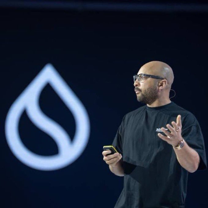 A bald man with glasses and a beard speaks on stage while holding a phone, standing in front of a large illuminated logo resembling a stylized 'S' or leaf.