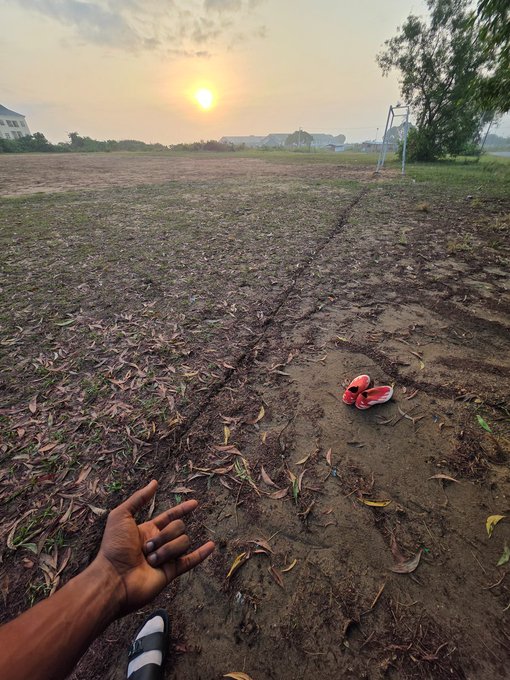 A sunset scene over an empty field with a pair of red and white sneakers abandoned on a dirt path, while a person's hand and sandaled foot are visible in the foreground.