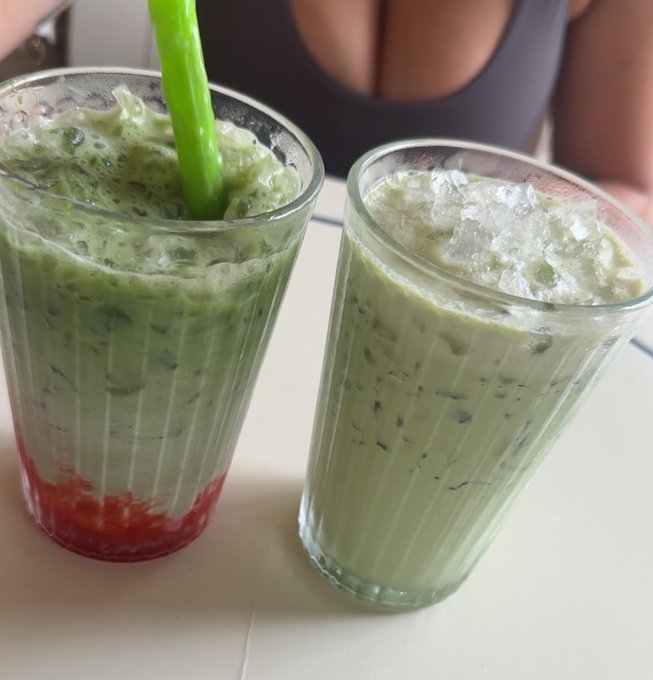 Two glasses of layered iced drinks, one with a green top and red bottom layer with a green straw, the other all green, placed on a white surface.
