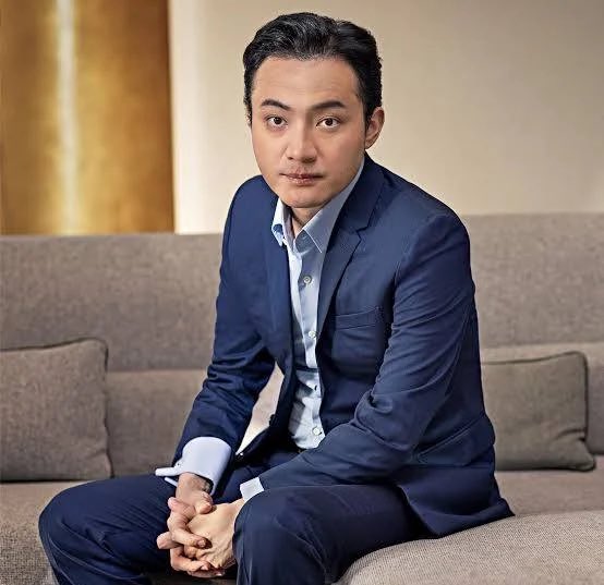 A professional portrait of a man in a navy blue suit and light blue shirt sitting on a gray couch.
