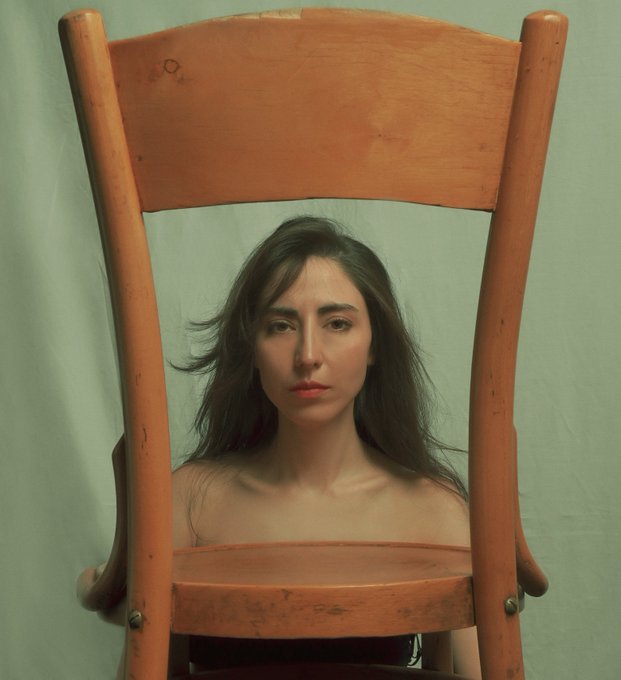 A woman with long dark hair and a serious expression sits behind a worn wooden chair against a pale green backdrop.