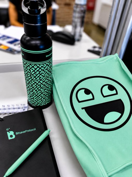 A promotional setup featuring a black water bottle with green patterns, a notebook with the 'BitsaveProtocol' logo, a pen, and a green fabric item with a smiley face design.