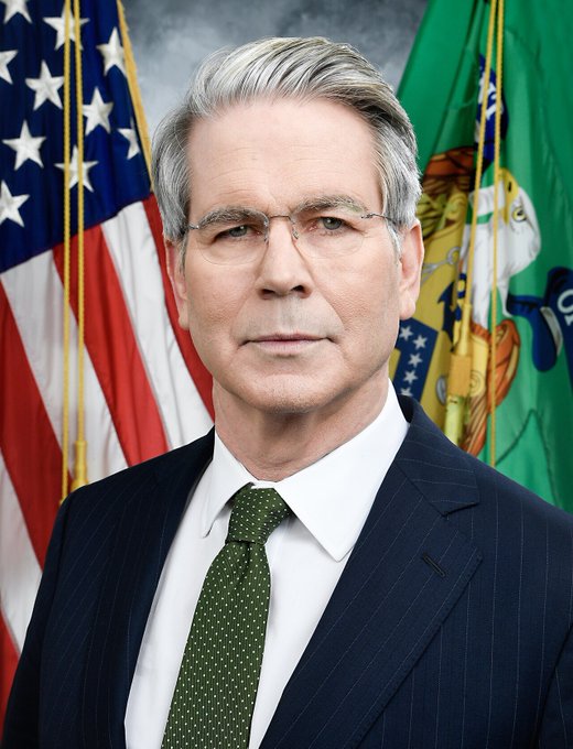 An official portrait of an older man with gray hair and glasses, wearing a dark pinstripe suit and a green patterned tie, standing in front of the United States flag and another official flag.