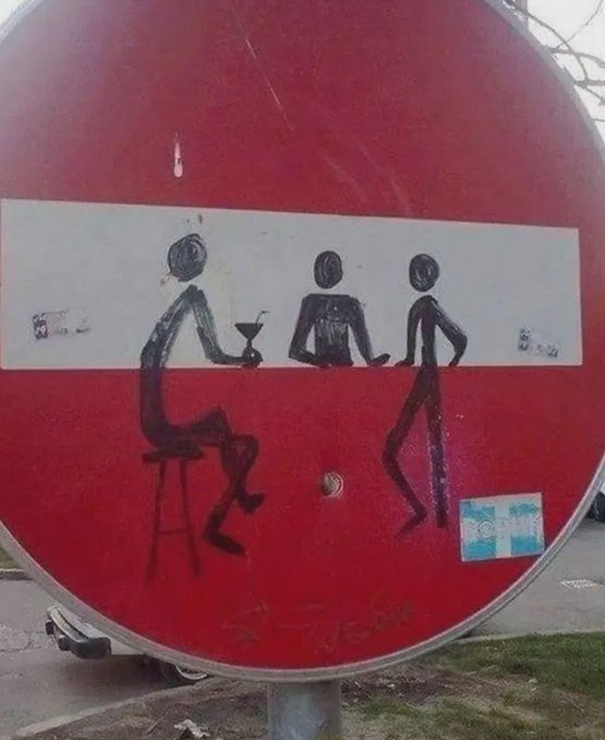 A red circular traffic sign that has been vandalized with graffiti depicting stick figures sitting and standing around a table drinking.