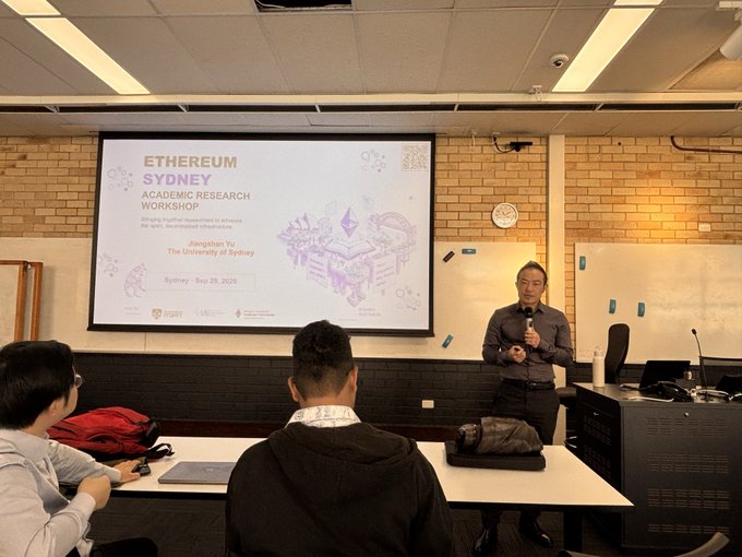 A man presenting at the 'Ethereum Sydney' academic research workshop hosted by The University of Sydney, standing in front of a projection screen with attendees seated at desks.
