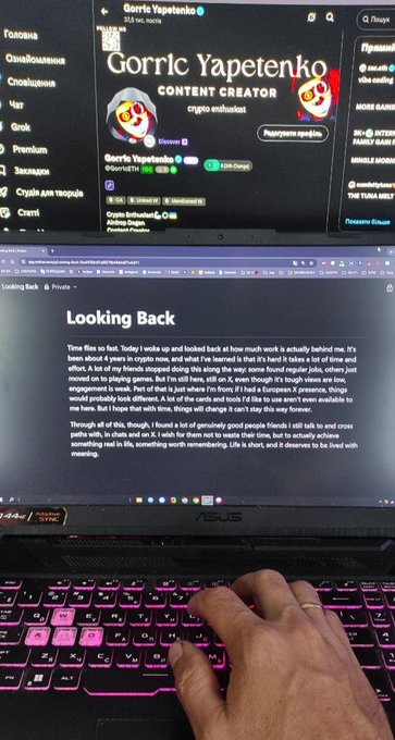 A person's hand types on a pink-backlit ASUS laptop showing a Twitch page for 'Gorrie Yapetenko' and a blog post titled 'Looking Back' about a creator's four years making crypto content.