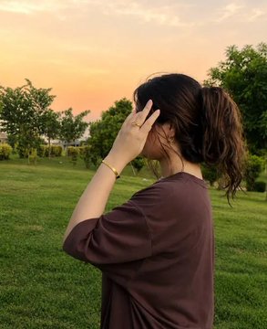 A woman with a ponytail standing in a park at sunset, shielding her eyes with her hand while looking into the distance.