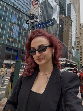 A woman with red hair, sunglasses, and a black blazer poses on a city street with a Madison Ave sign visible in the background.