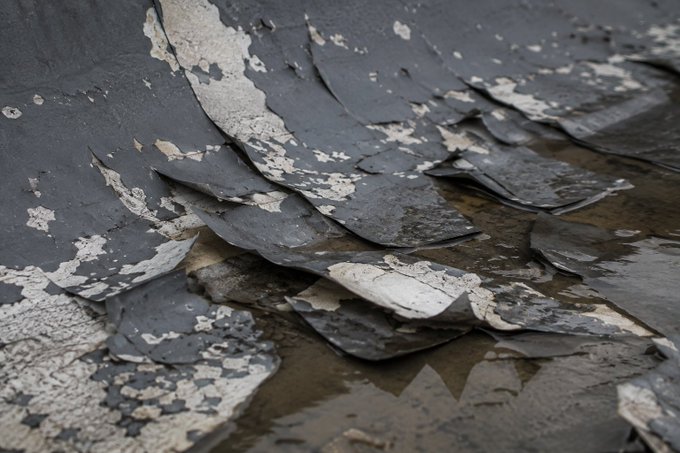 A textured, rocky surface featuring dark, peeling layers of rock or paint that reveal lighter gray and tan colors underneath.