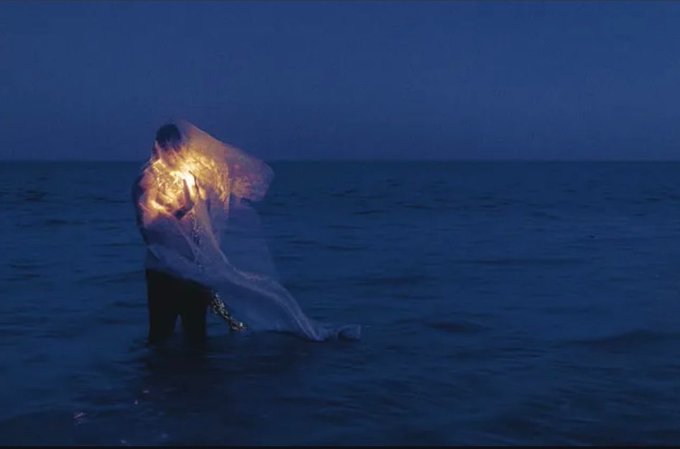 A person standing in dark blue ocean water at dusk, holding up a glowing, translucent white fabric illuminated from within by warm yellow light.