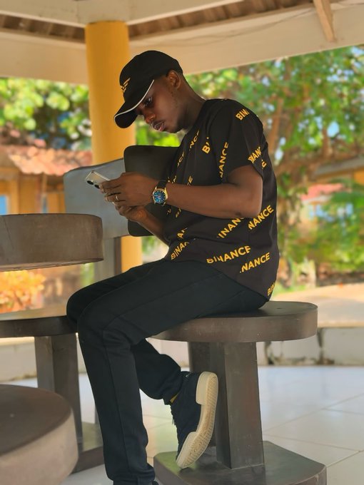 A young man wearing a black cap and a 'BINANCE' branded shirt sits on a wooden bench, looking down at his phone.