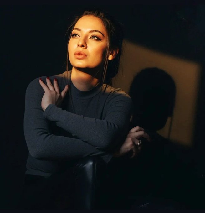 A moody, low-light portrait of a woman in a dark long-sleeved top gazing upward with dramatic side lighting.