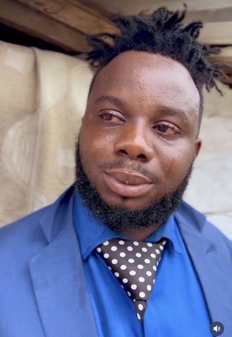 A close-up portrait of a man with short dreadlocks and a beard, wearing a blue shirt, blue blazer, and a black polka-dot tie.