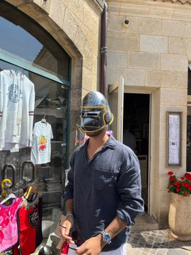 A man wearing a gold/chrome motorcycle helmet and sunglasses stands on a sunny street in front of a stone building with a shop window displaying clothing.