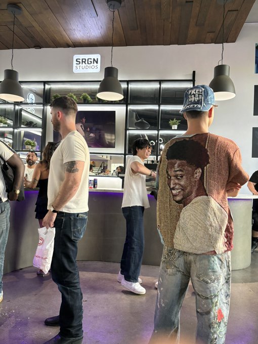 People gather at an indoor event space with a 'SRGN STUDIOS' sign overhead; one person wears a brown shirt featuring a large printed face graphic on the back, along with distressed jeans and a baseball cap.