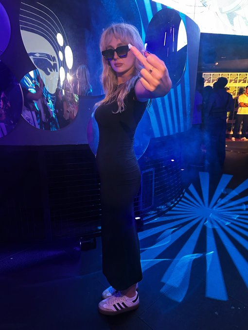 A blonde woman in a black dress and sunglasses posing at a dimly lit, blue-toned venue with futuristic decor.
